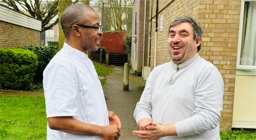 Two men converse outside beside a brick building and green lawn.
