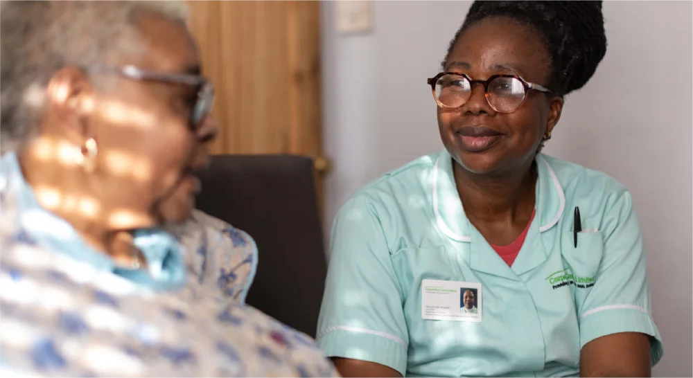 Nurse in green uniform talks with an elderly patient indoors.