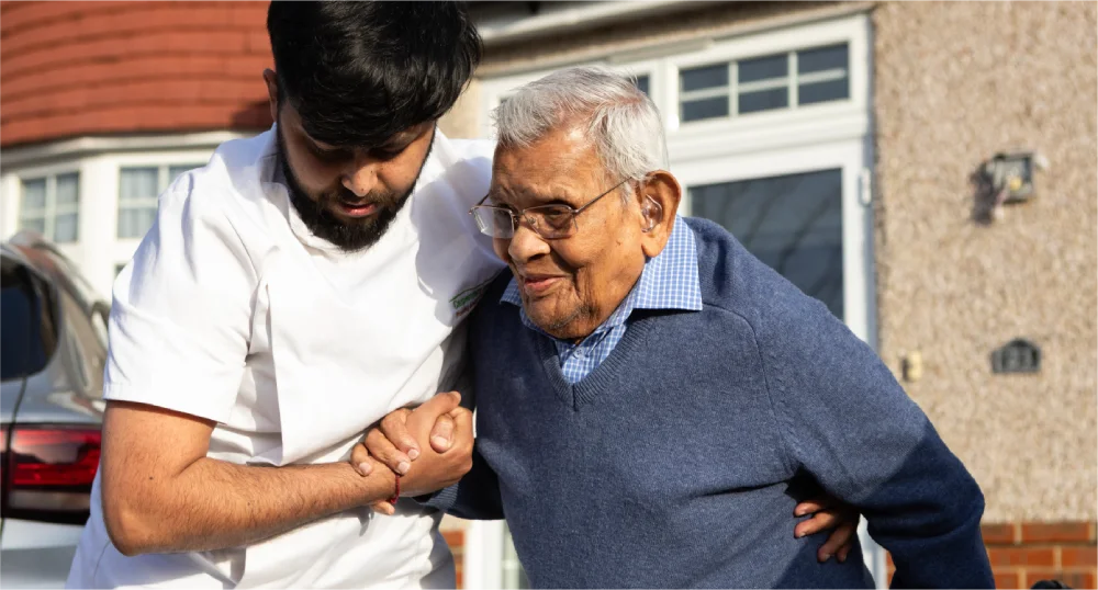Young caregiver assists an elderly man walking outside a house.