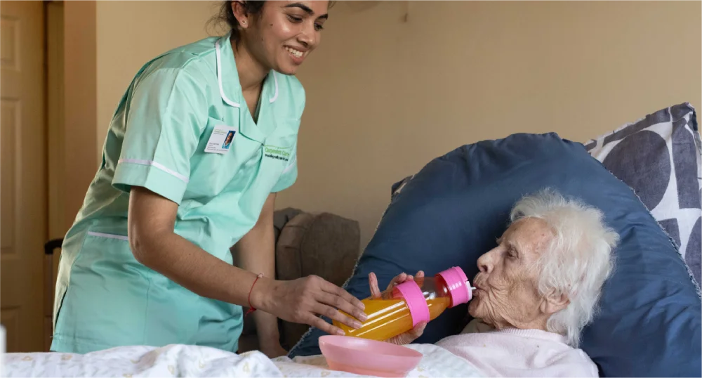 Nurse helps an elderly patient drink from a bottle in bed.